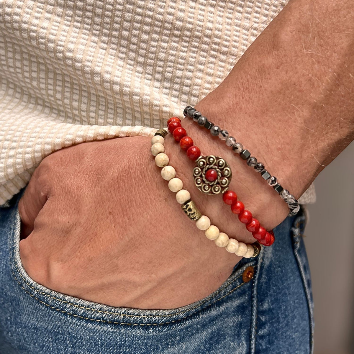 Un hand en un jean pocket wears the JAREDJAMIN Joyería Online Red Barron pulsera Set: audaz red Coral con metal centerpiece, cream, black-and-white marbled, y red con skull bead. Paired con a cream texturizado shirt.