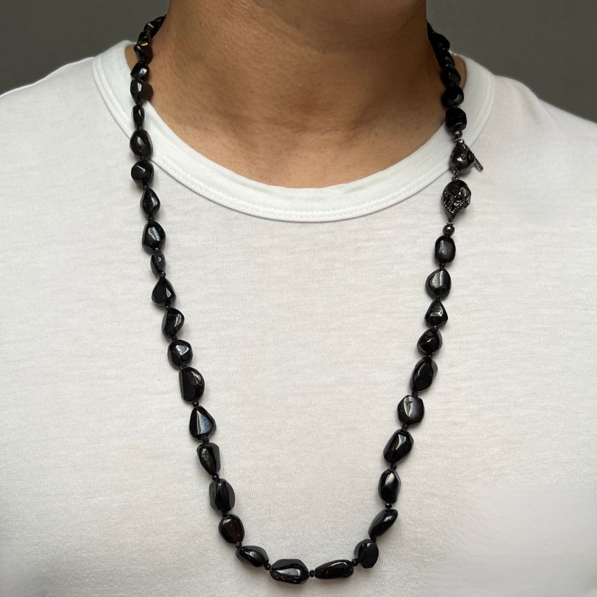 A person is shown from the shoulders up wearing a white crew-neck shirt and the JAREDJAMIN Jewelry Online Rocks of Sangria Skull-Clasp Necklace, featuring black irregular-shaped spinel beads, a small silver pendant, and a unique skull clasp.