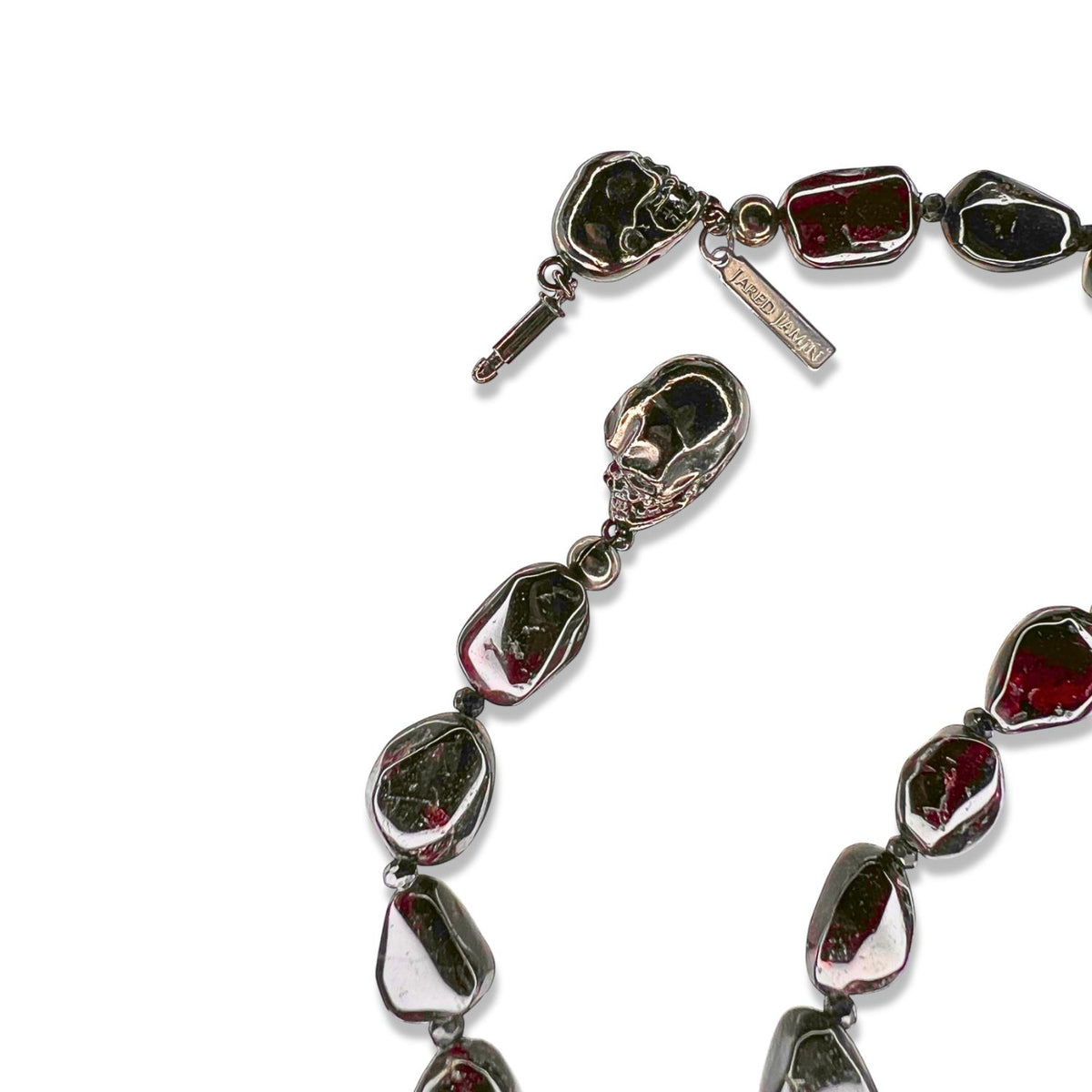 A close-up of JAREDJAMIN Jewelry Online&#39;s Rocks of Sangria Skull-Clasp Necklace features glossy, dark red garnet and black spinel pebbles linked by metal loops, a double-skull clasp, and a branded rectangular tag on a white background.