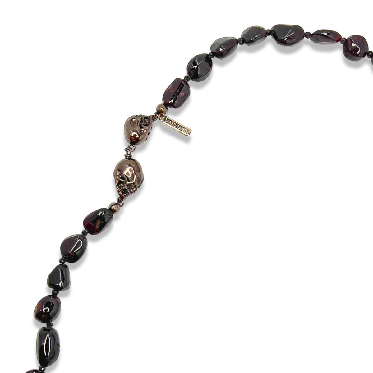A クローズアップ of JAREDJAMIN ジュエリーオンライン Rocks of Sangria Skull-Clasp ネックレス shows pebble-shaped garnets, dark stones, a metallic skull チャーム, small beads,とa "LIBERTY FAIRS" tag, arranged の中の gentle curve on ホワイト.