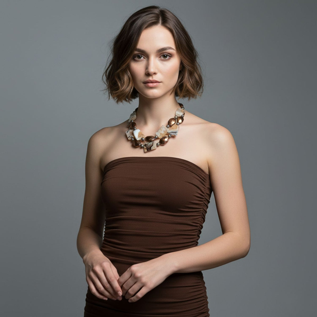 A young woman with short, wavy brown hair wears a strapless brown dress and the Rocky Mountain Necklace by JAREDJAMIN Jewelry Online, standing against a plain gray background with her hands lightly clasped in front of her.