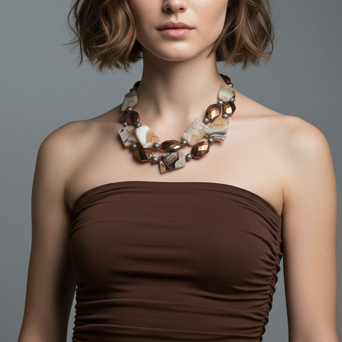 A woman with short, wavy brown hair wears a strapless, ruched brown dress and the JAREDJAMIN Jewelry Online Rocky Mountain Necklace, featuring large agate stones. A plain gray background highlights her outfit and statement jewelry.