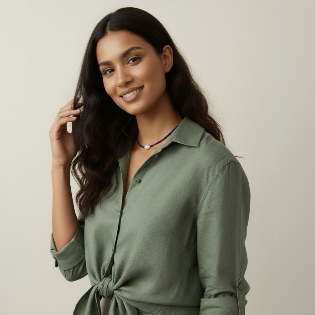 A woman with long, wavy dark hair smiles gently, wearing an olive green button-up shirt tied at the waist and the Rowan Necklace Lapis Lazuli by JAREDJAMIN Jewelry Online. Her hand is raised to her hair against a plain, light background.