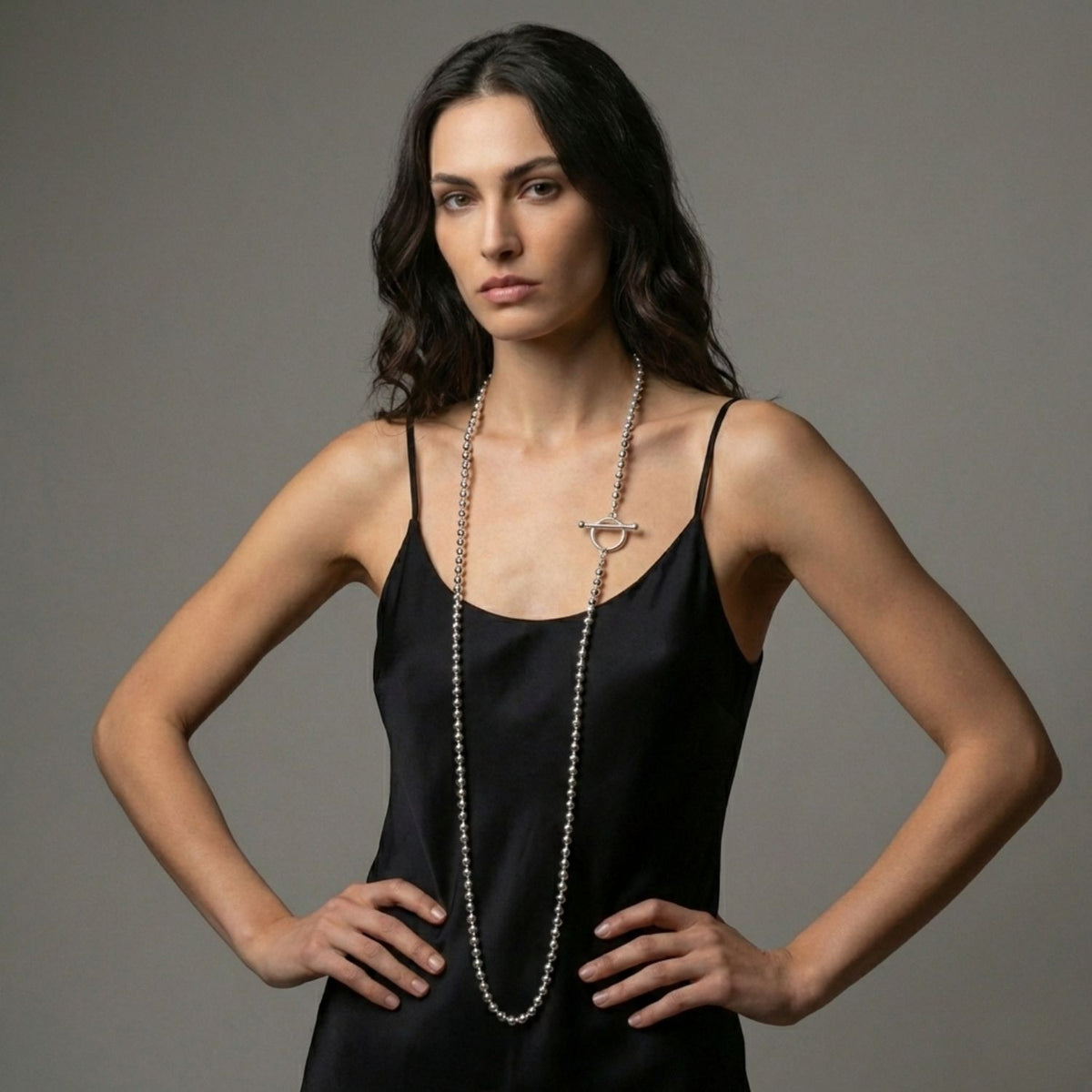 A woman with wavy dark hair stands hands on hips in a sleeveless black dress, wearing the Silver Constellation Orbita Necklace by JAREDJAMIN Jewelry Online—a reflective silver piece with a circular clasp—against a plain gray background.