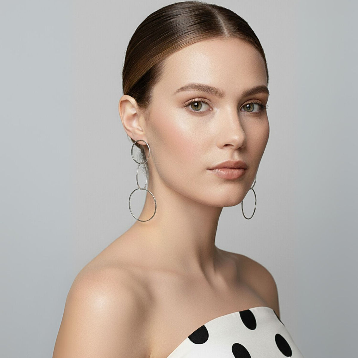 A woman with light skin and straight brown hair in a low bun wears JAREDJAMIN Jewelry Online’s Silver Lindy Links Post Earrings and a strapless white top with black polka dots, standing against a plain light gray background with a neutral expression.