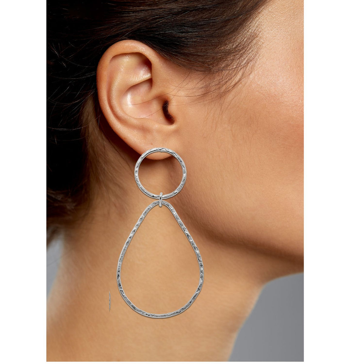 A close-up shows a person wearing Silver Textured Tear Post Earrings by JAREDJAMIN Jewelry Online. The large silver earrings feature a circular top and pear-shaped teardrop bottom with a hammered texture, attached to the earlobe. Hair is pulled back against a neutral gray background, highlighting the elegant design.