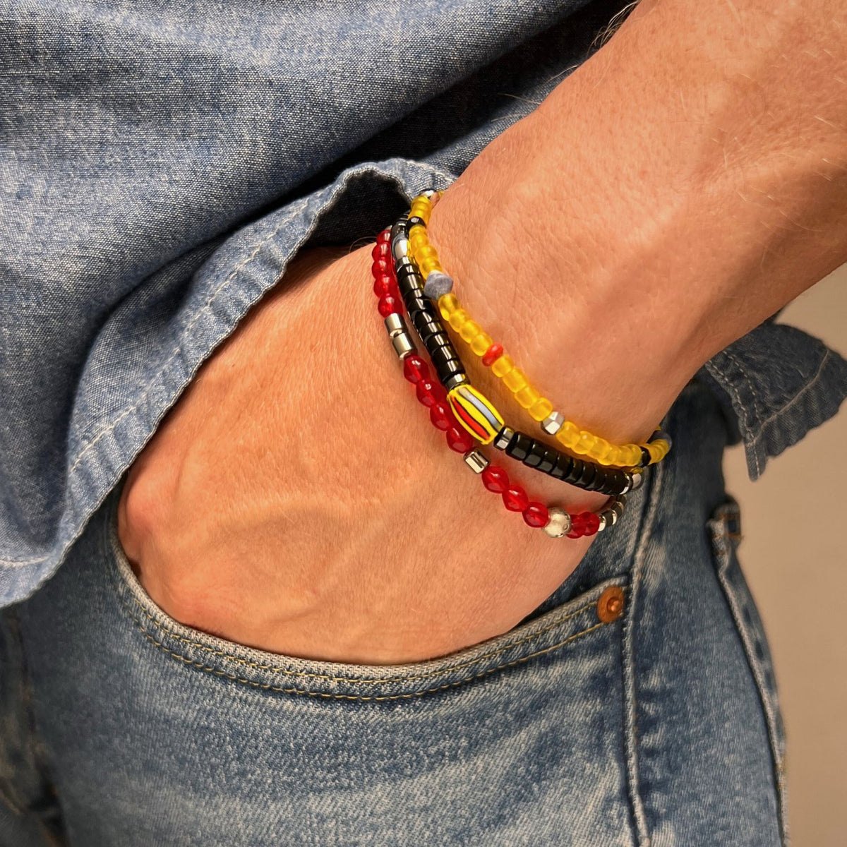 A クローズアップ shows a hand in ブルー jeans sporting Samurai ブレスレット Set JAREDJAMIN製 ジュエリーオンライン—layered stretch ブレスレットとyellow, レッド, ブラック, シルバー beads,とhematite. A rolled-カフ ブルー denim shirt completes look.