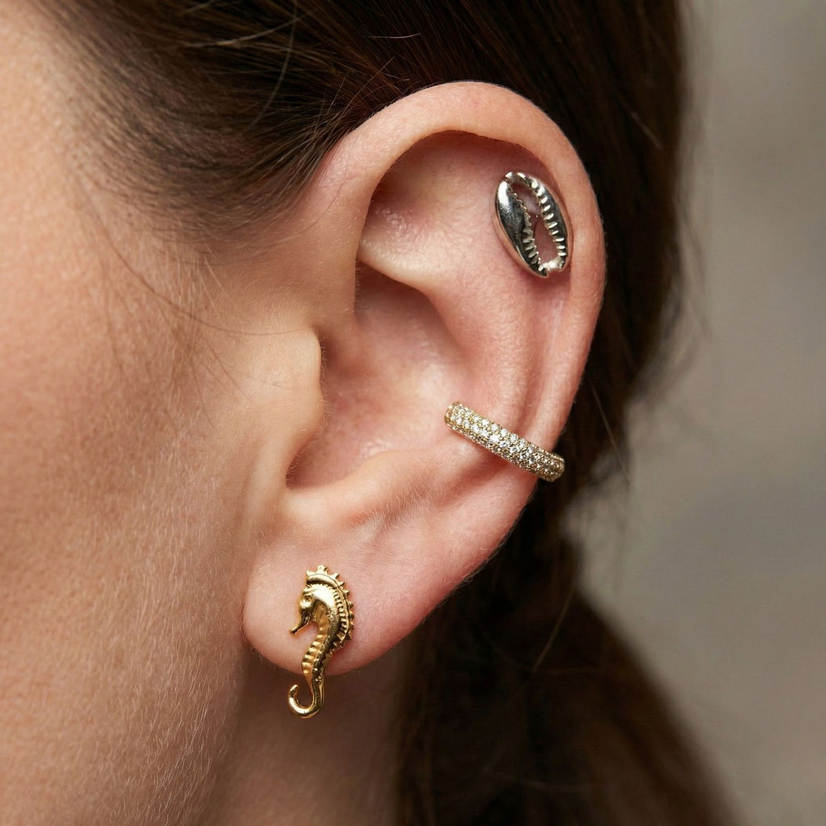 Un primer plano shows a person's ear adorned con JAREDJAMIN Joyería Online's plata Cowrie pendiente de botón Post pendientes, plus other coastal-inspired pieces. Brown hair is pulled back to highlight the plata pendiente de botón y accompanying joyería.