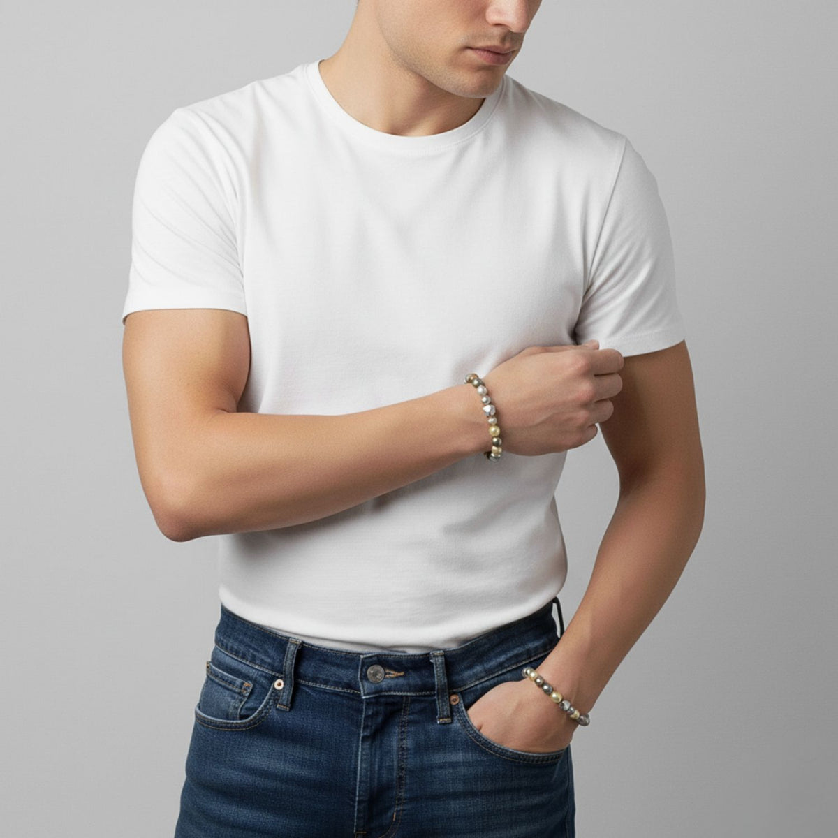A man in a plain white t-shirt and blue jeans stands against a gray background, wearing matching Seabed Pebble Stretch Bracelets from JAREDJAMIN Jewelry Online on his left wrist. One bracelet features freshwater pearls; he looks downward, face partially out of frame.