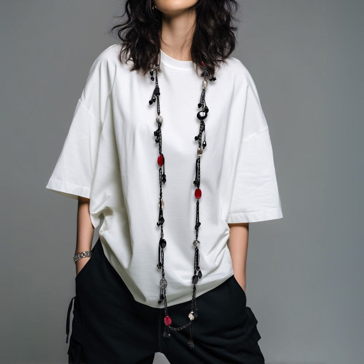 A woman with wavy dark hair wears an oversized white tee, loose black pants, and the See You Saloon Necklace by JAREDJAMIN Jewelry Online. She stands against a plain gray background, hands in pockets, her face partially out of frame.