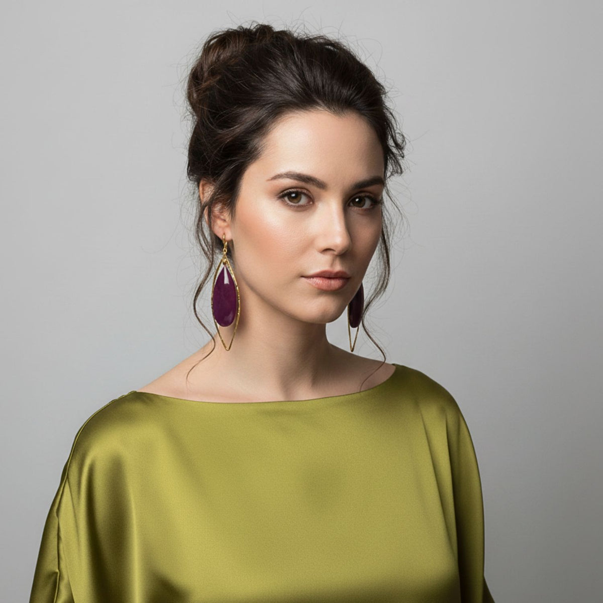 A young woman with dark, wavy hair in a loose updo wears a silky olive top and Seraphine Plum Drop Leverback Earrings by JAREDJAMIN Jewelry Online, sporting natural makeup and posing calmly against a plain light gray background.