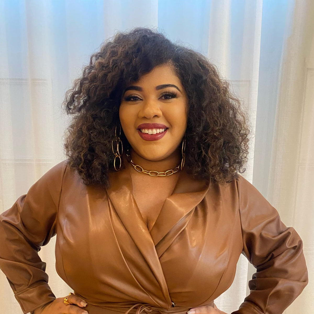 A curly brown-haired woman smiles, wearing a brown leather-style wrap dress, Shackleford Chandelier Post Earrings by JAREDJAMIN Jewelry Online, and an 18k gold chain necklace. Her hands rest on her hips against a backdrop of light-colored curtains for a soft and elegant ambiance.