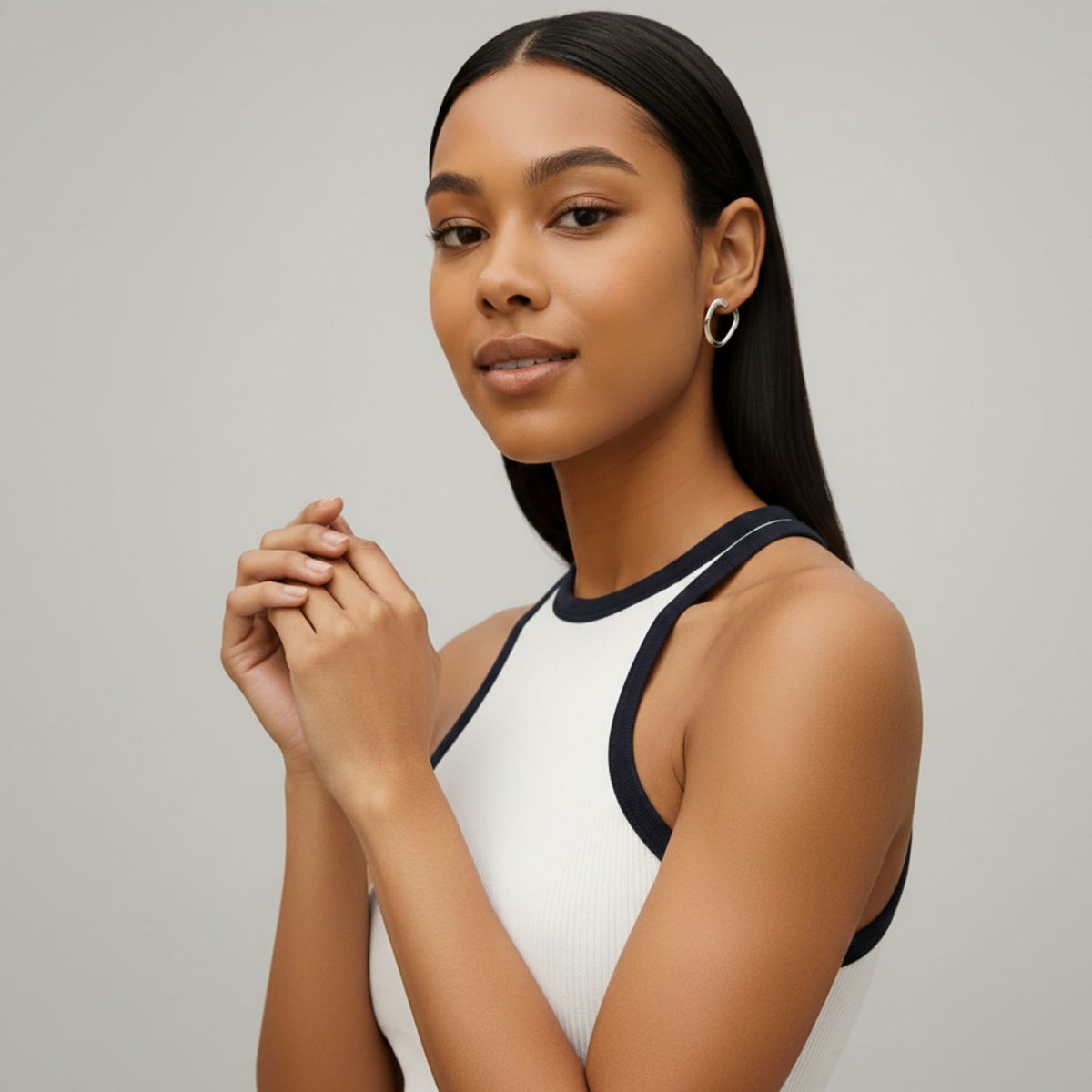 Un woman con straight, dark hair parted in the middle wears a sleeveless white top con black trim y plata Billie pendiente de botón Post pendientes de JAREDJAMIN Joyería Online. She clasps her hands y looks confidently at the camera against a light gray background.