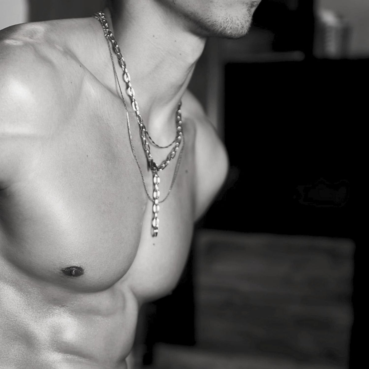 Black-and-white close-up of a shirtless upper torso highlights defined muscles and standout skin texture, featuring the JAREDJAMIN Jewelry Online Silver Chain Reaction Necklace layered with another necklace; background is blurred.