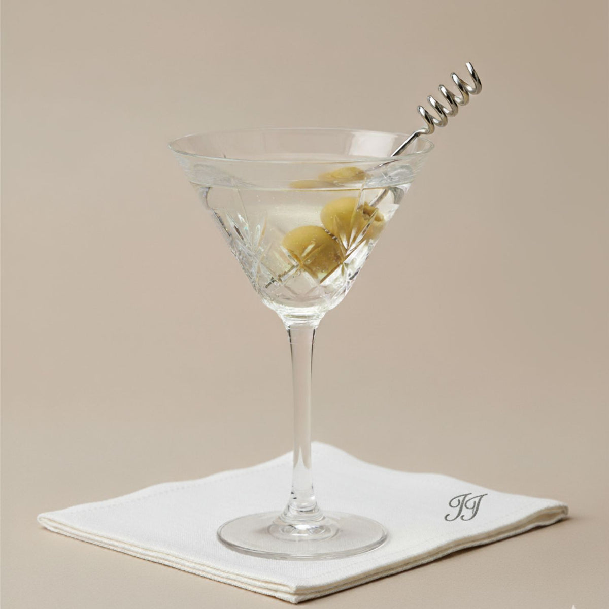 A classic martini with three green olives on JAREDJAMIN Barware and More Silver Corkscrew Cocktail Skewers, served in a faceted glass atop a white napkin embroidered with &quot;JJ&quot; on a neutral beige background.