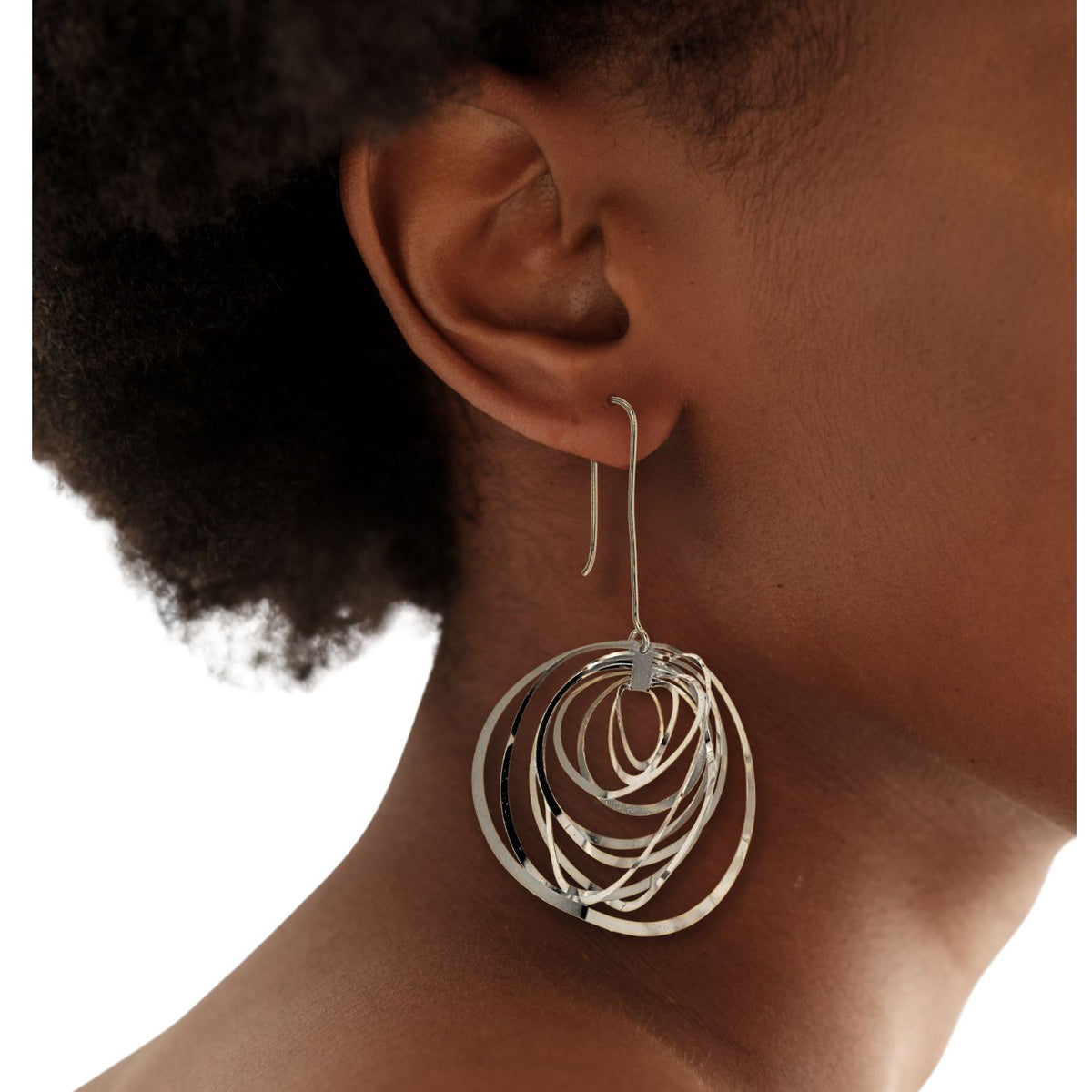 Someone with short curly hair sports a large Silver Dahlia Hook Earring by JAREDJAMIN Jewelry Online, featuring artistic concentric loops hanging elegantly from a French hook. A plain white background accentuates the earring&#39;s intricate design.