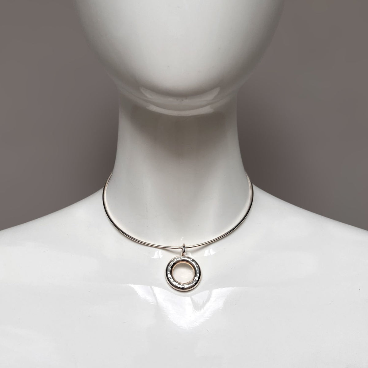 A glossy white mannequin displays the JAREDJAMIN Jewelry Online Silver Doughnut Pendant Collar Necklace, featuring a smooth ring encasing a textured circle. It fastens with a lobster clasp, set against a neutral gray background.