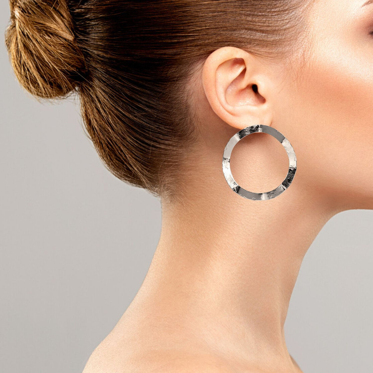 Un woman con light skin y brown hair en un bun wears JAREDJAMIN Joyería Online's plata Duet Post pendientes. Los large, texturizado, tarnish-free plata plated pendientes stand out against her minimal makeup y a plain light gray background.