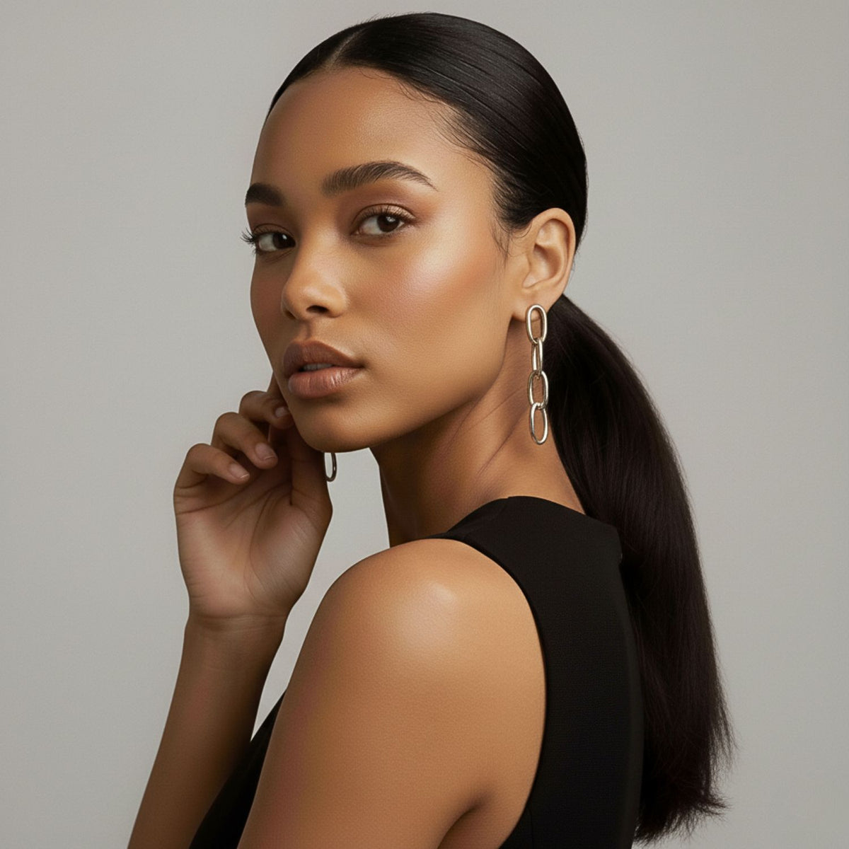 Un woman con smooth, medium brown skin y long, dark hair en un low ponytail poses in profile wearing a sleeveless black top y plata Everly colgante cadena Post pendientes de JAREDJAMIN Joyería Online. Her makeup is natural as she gently touches her face.