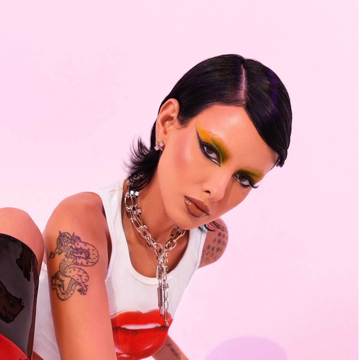 Un person con slick, short black hair poses against a pink background, wearing audaz yellow-green eyeshadow y glossy lips. They have a dragon tattoo y wear the plata Everly Every-Other collar de JAREDJAMIN Joyería Online con a red lips print white tank top.
