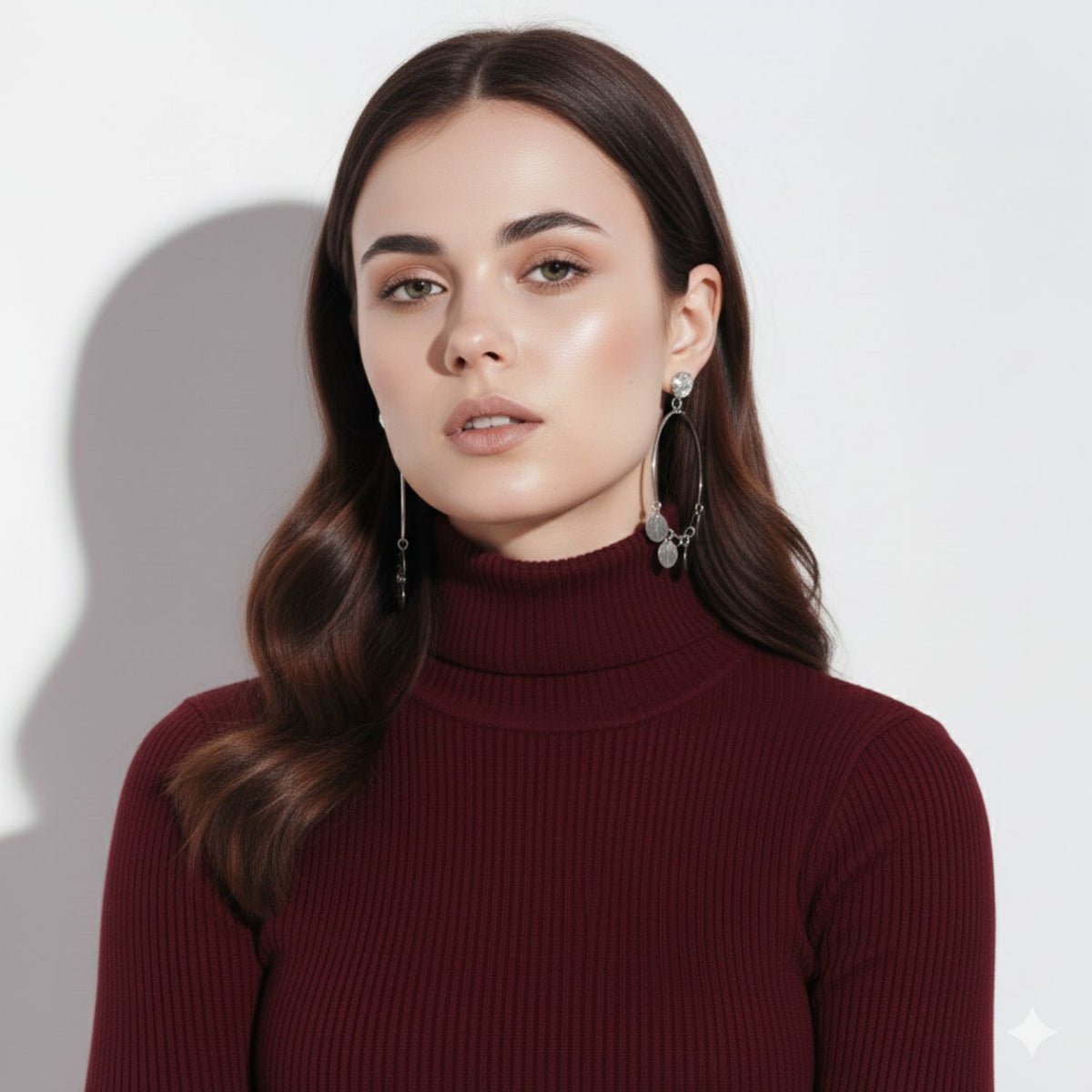Un woman con wavy brown hair wears a burgundy turtleneck y plata Fortuna Chandelier pendientes de JAREDJAMIN Joyería Online. She looks confidently at the camera against a fondo blanco, con soft natural makeup y a neutral expression.