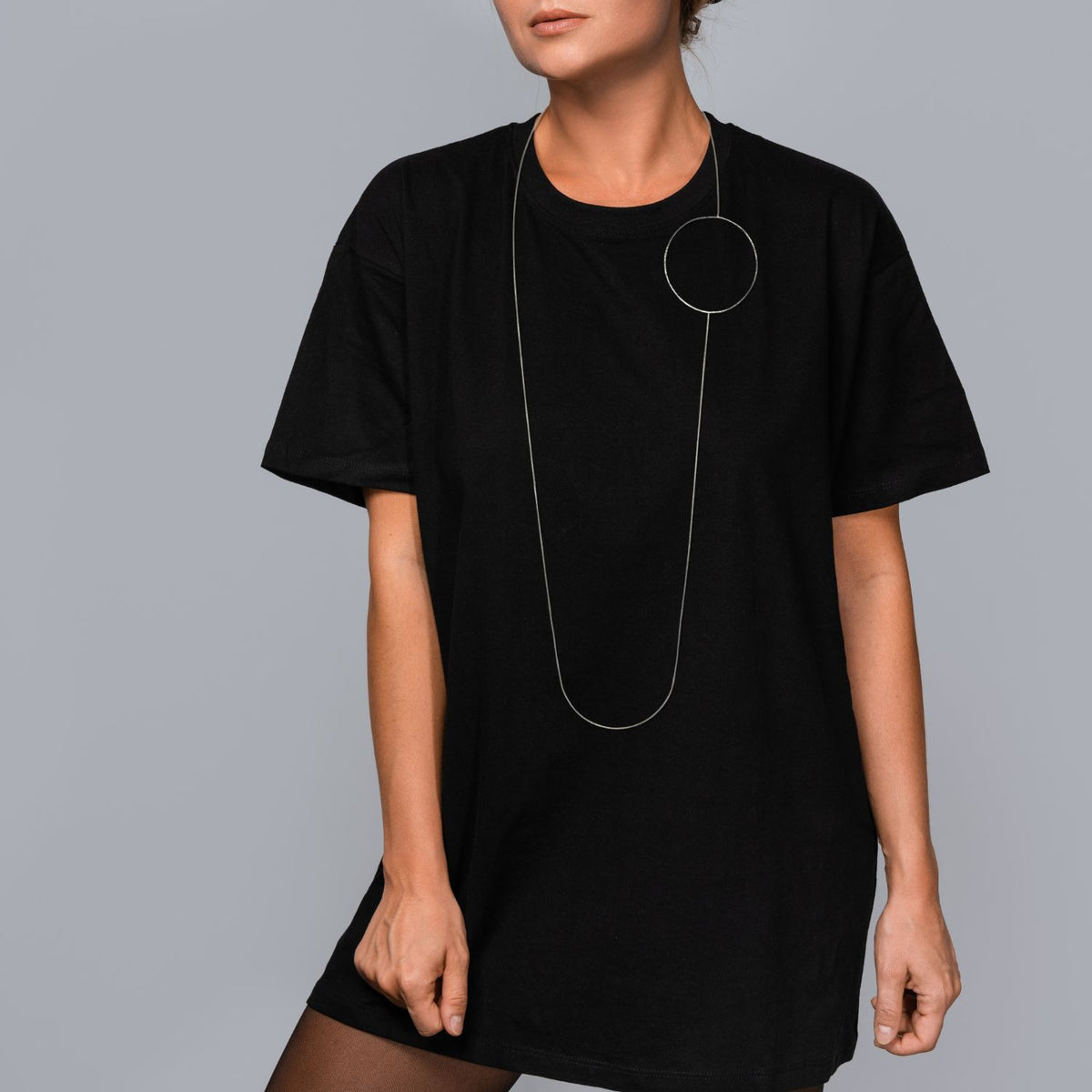 Against a gray background, a person in a loose black t-shirt and sheer tights wears the Silver Halo Necklace by JAREDJAMIN Jewelry Online, featuring a large circular pendant and toggle clasp resting on their chest.