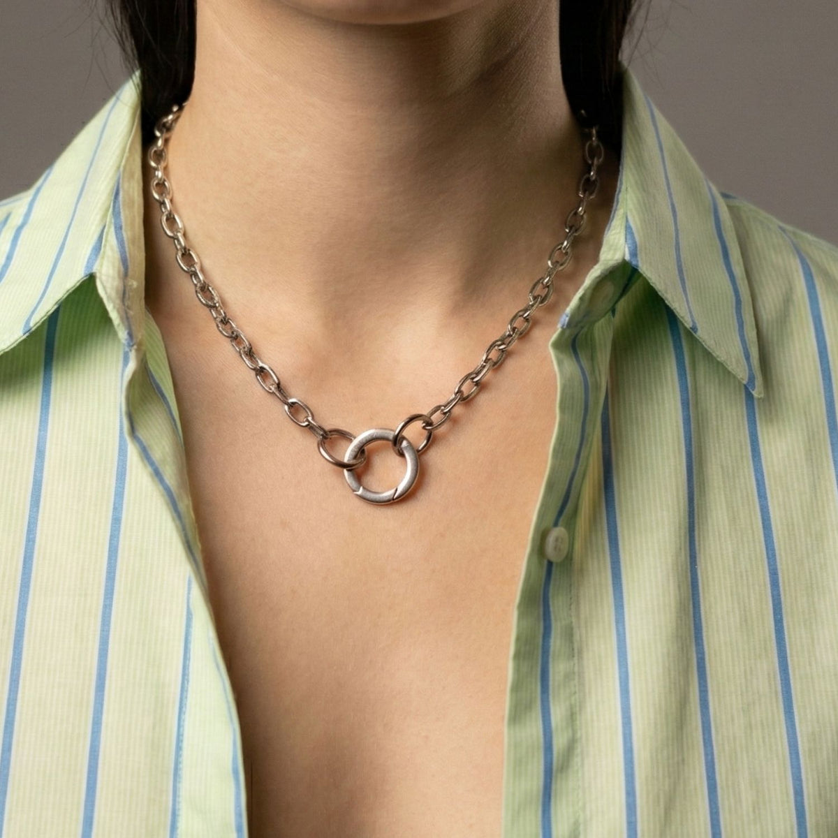 A person in a green and blue striped shirt with an open collar wears a chunky silver chain necklace featuring three interlocking rings and the Silver Medium Circular Clicker Clasp from JAREDJAMIN Jewelry Online. Face not visible; plain, neutral background.