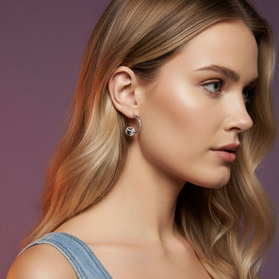 A young woman with long, wavy blonde hair and glowing skin is shown in profile against a purple background, wearing a sleeveless denim top and JAREDJAMIN Jewelry Online’s Silver Orbita-U Post Earrings. Her makeup is natural and her expression calm.