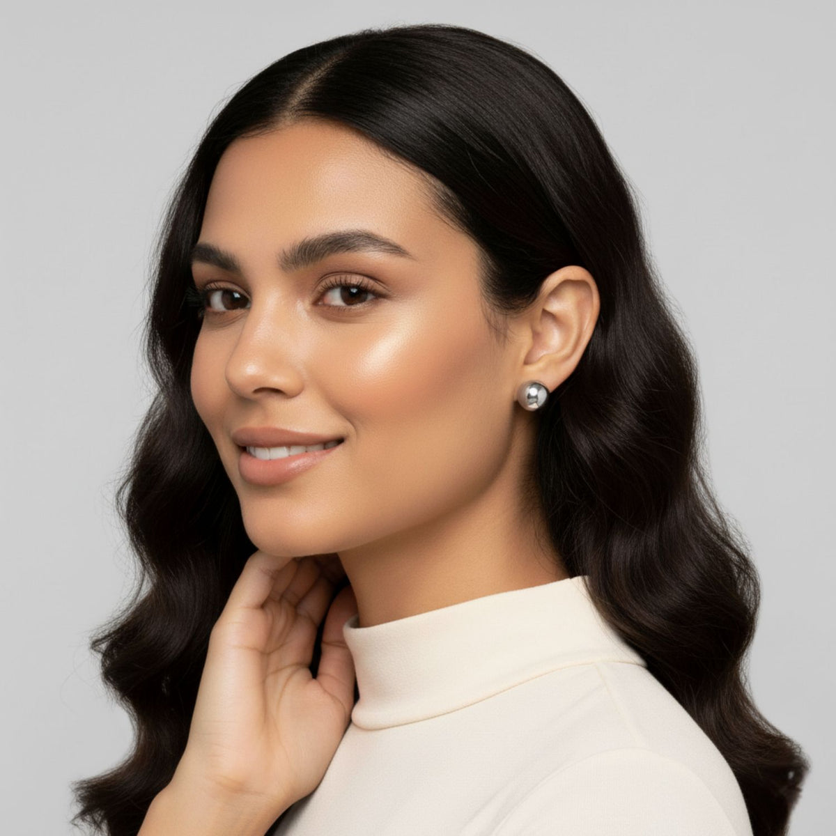 Un woman con long, wavy dark hair y glowing skin smiles softly. She wears JAREDJAMIN Joyería Online's plata Orbita pendiente de botón Post pendientes y a cream turtleneck top, her hand gently touching her neck against a plain light gray background.