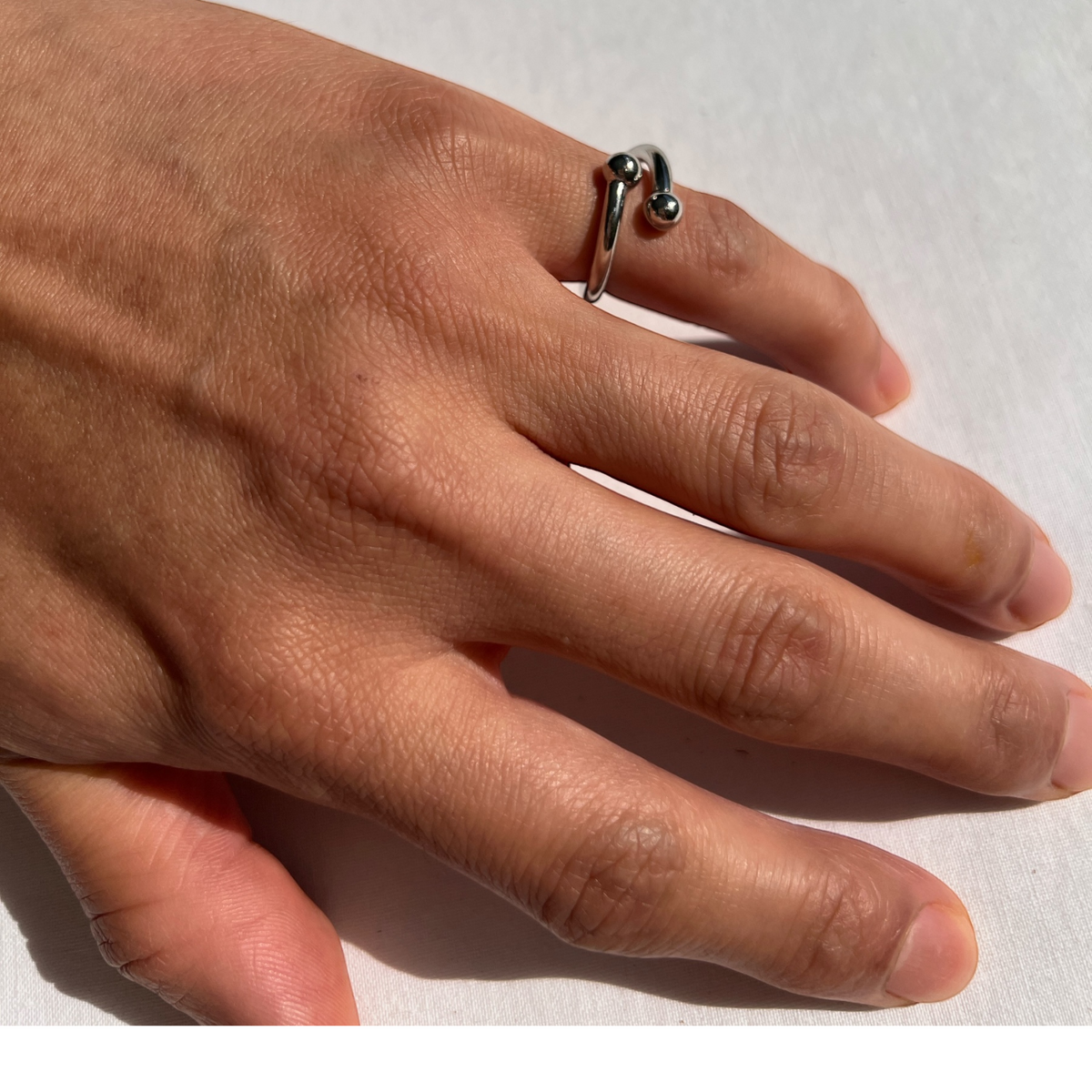 A hand rests on a white surface, wearing the tarnish-proof Over Easy Orbita Ring by JAREDJAMIN Jewelry Online. The elegant loop design with two small spheres enhances the finger, as natural skin tone and sunlight highlight the ring&#39;s sheen, casting soft shadows.