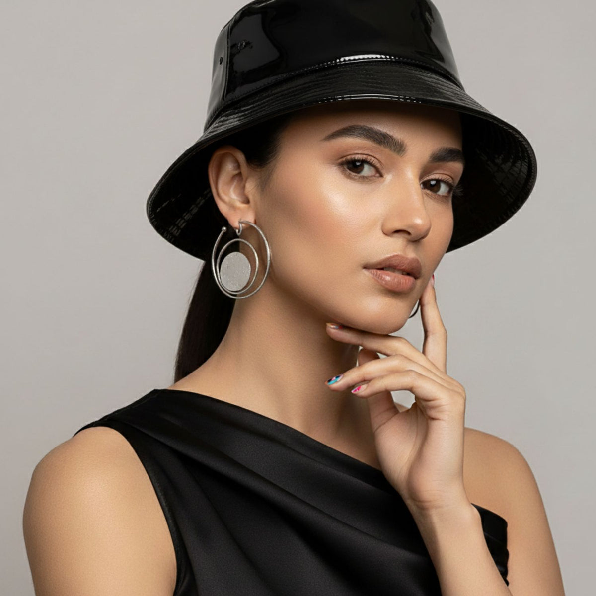 Un woman dans un black vinyl bucket hat et asymmetrical top poses avec her hand to her chin, wearing straight dark hair, subtle makeup, multicolored nails, et JAREDJAMIN Bijoux en Ligne’s argent Peacock Clip On créole boucles d'oreilles.