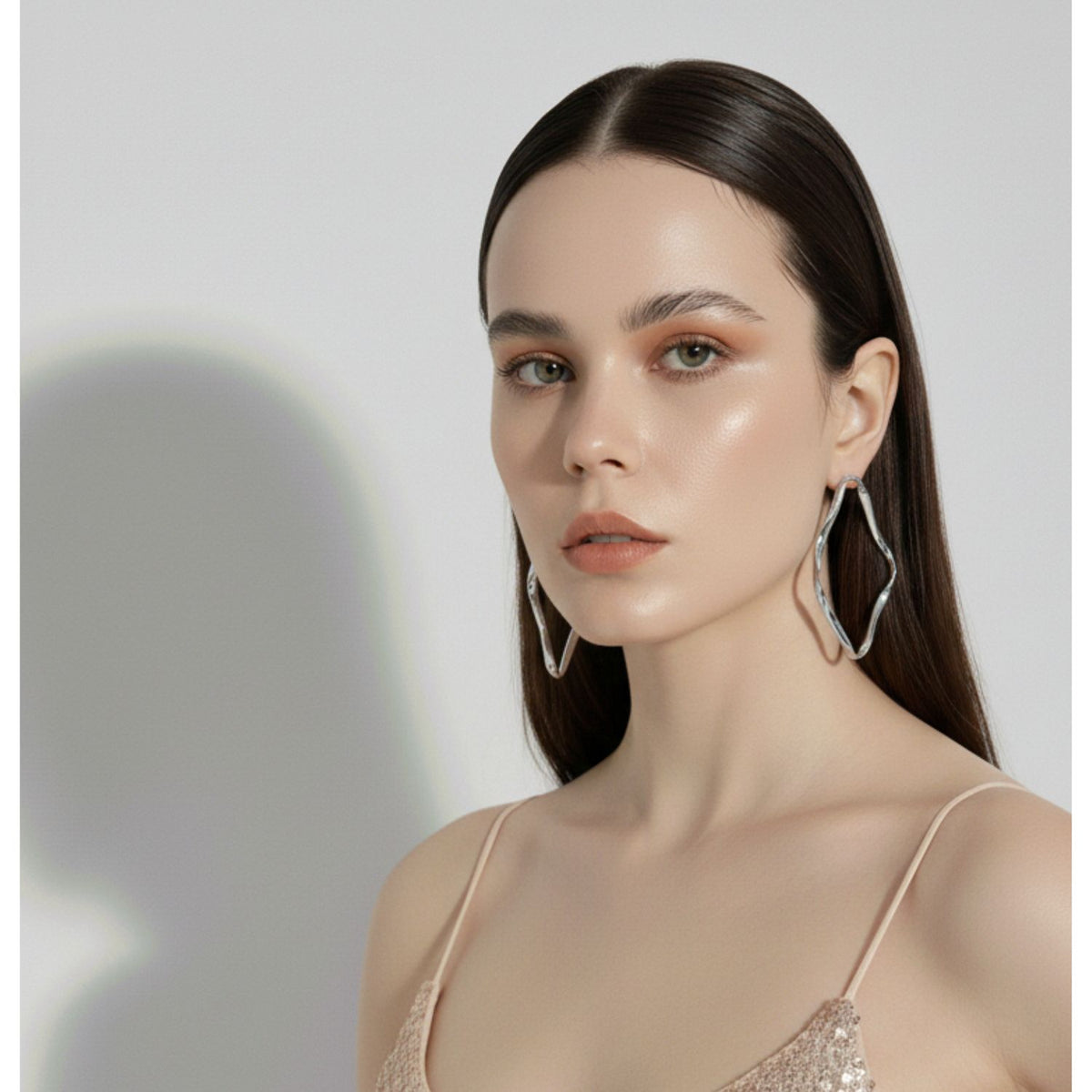 A woman with straight, dark brown hair wears JAREDJAMIN Jewelry Online&#39;s Silver Ribbonelle Earrings and a light sequin spaghetti-strap top. Her dewy skin and natural makeup stand out as she gazes at the camera against a plain light background.