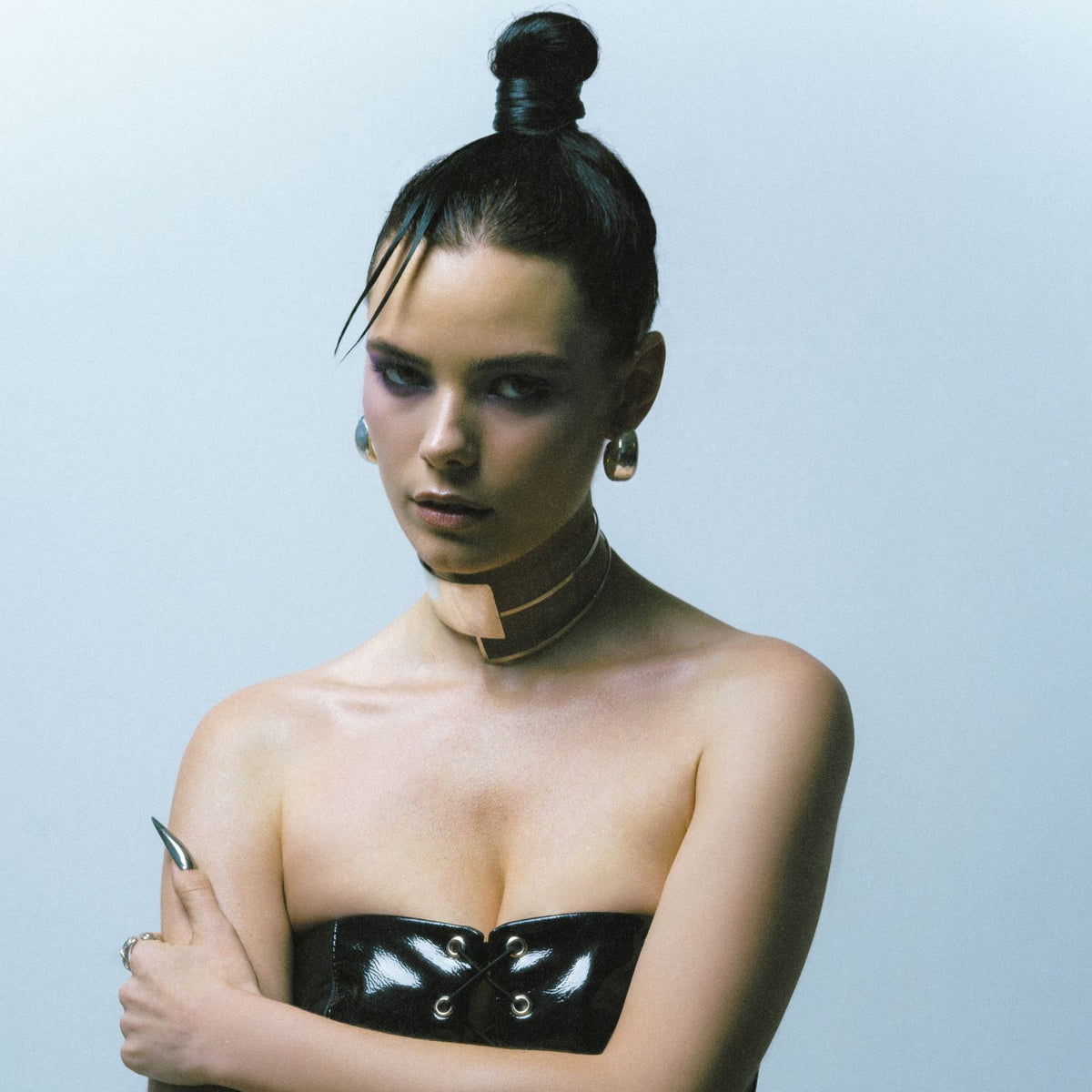 Un woman avec dark hair dans un topknot et audacieux makeup poses against a plain background, wearing JAREDJAMIN Bijoux en Ligne’s argent Rush créole boucles d'oreilles, a brillant black strapless top, a wide beige choker, et long metallic nails.