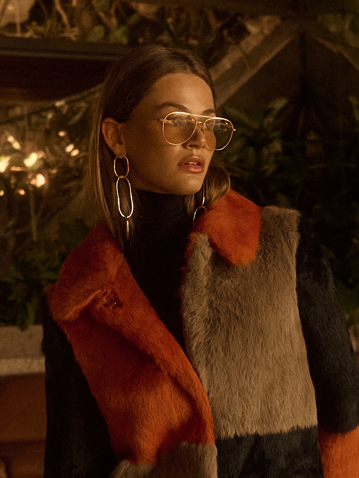 A person in aviator glasses and Shackle Link Dangle Post Earrings from JAREDJAMIN Jewelry Online is styled in a chic faux fur coat with orange, gray, and black patches against a softly lit background with warm hues, blurred plants, and a cozy ambiance.