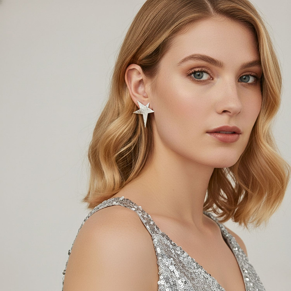Un woman con shoulder-length blonde hair wears a plata sequin dress y JAREDJAMIN Joyería Online's plata Shooting Star pendiente de botón Post pendientes. She gazes to the side con a neutral expression y subtle, natural makeup against a light background.