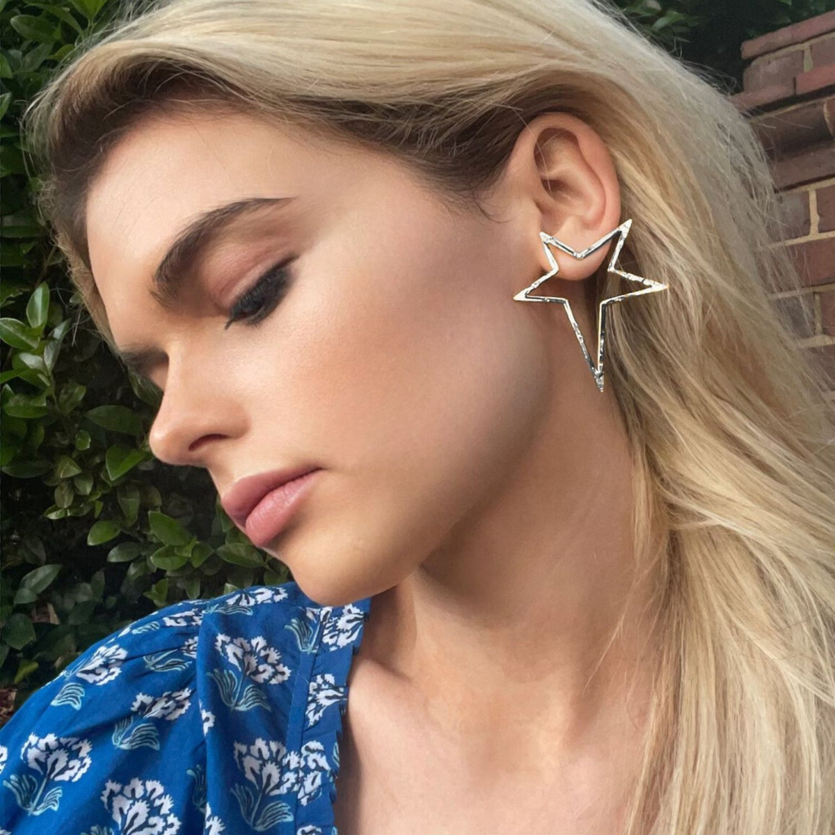 A person with blonde hair and soft makeup wears JAREDJAMIN Jewelry Online&#39;s Silver Shooting Star Dust Post Earrings, eyes closed as they look down. The green foliage and brick wall backdrop contrasts with their blue patterned top.