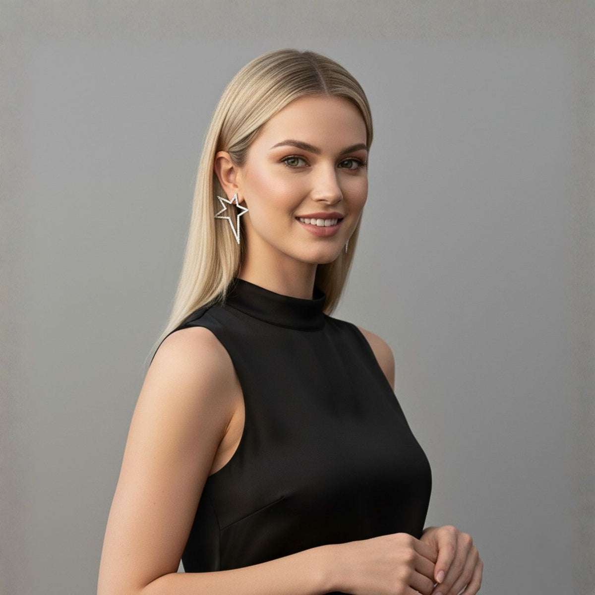 Un woman con straight blonde hair y light skin smiles gently, wearing a sleeveless black high-neck top y JAREDJAMIN Joyería Online's plata Shooting Star Dust Post pendientes. Los plain gray background adds a modern, minimalistic touch.