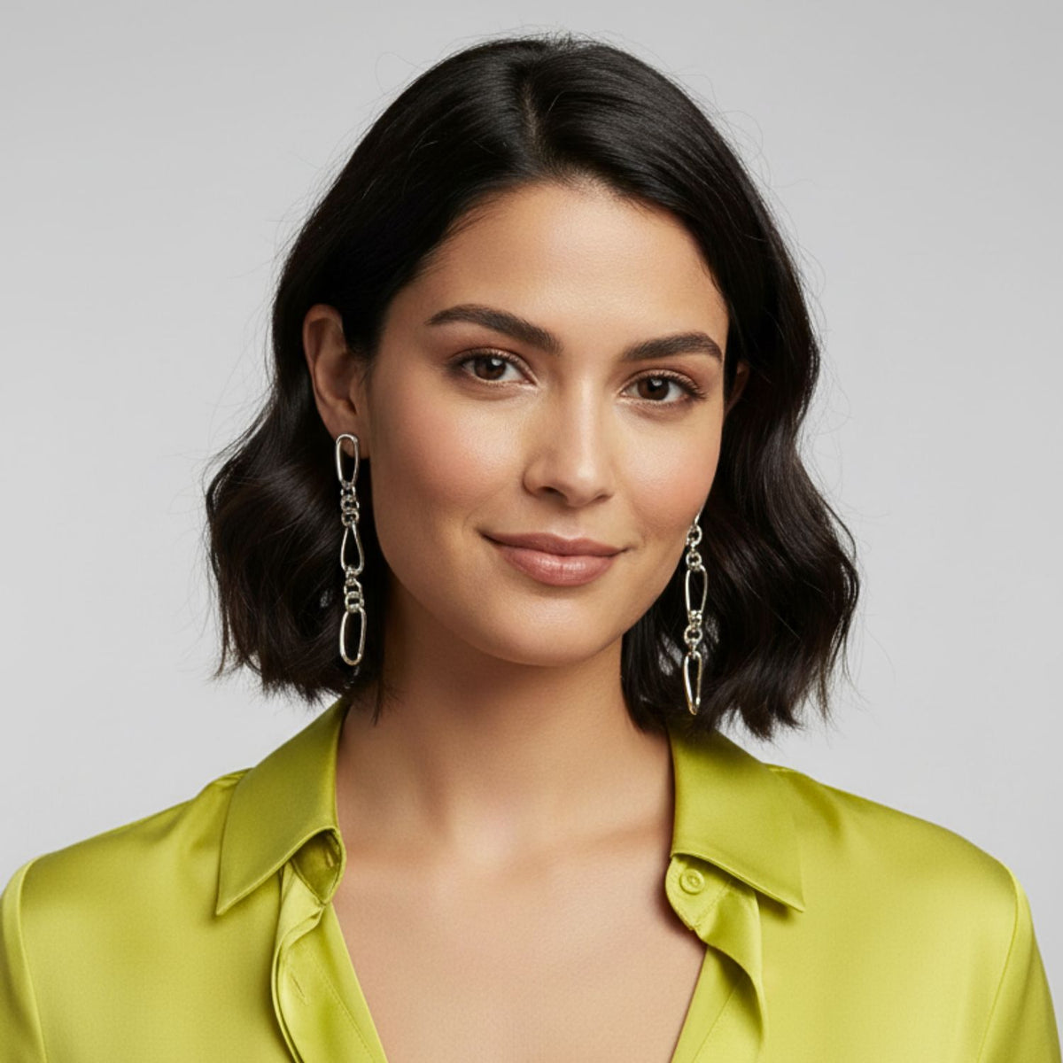 A womanとwavy, chin-length dark hair smiles softly, wearing JAREDJAMIN ジュエリーオンライン シルバー Skyler チェーン Post イヤリングとa 光沢のある light グリーン satin blouse. A plain light gray background highlights her エレガント, 磨き上げた look.