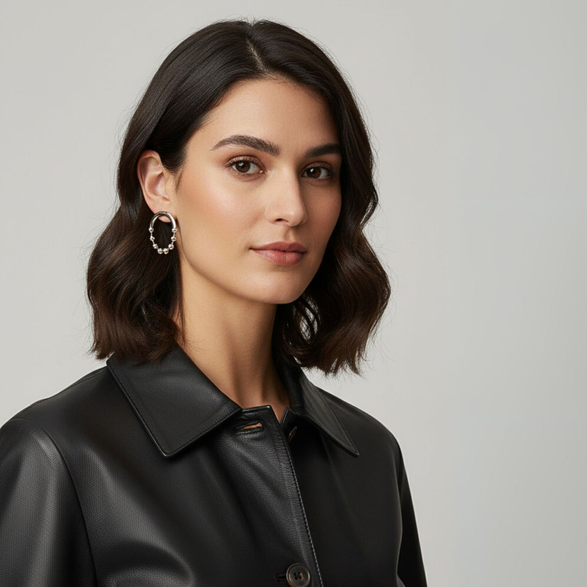 Un woman con shoulder-length dark wavy hair wears a black leather collared jacket y plata Small Orbita Amari Post pendientes de JAREDJAMIN Joyería Online, standing against a plain light gray background y gazing calmly at the camera.