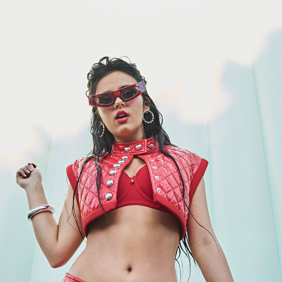 A woman in a red quilted crop top and bikini poses confidently, accessorized with pink cat-eye sunglasses, Silver Small Orbita Amari Post Earrings from JAREDJAMIN Jewelry Online, and an 18K Gold Plate bracelet. Her wet hair flows against a soft, light blue background reminiscent of daylight.