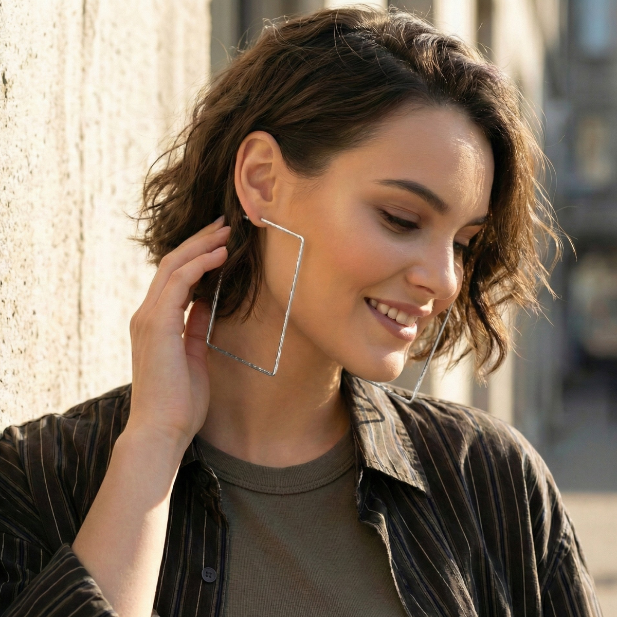 A woman with short, wavy brown hair smiles, touching JAREDJAMIN Jewelry Online’s Silver Squaretastic Hoop Post Earrings. She stands by a textured concrete wall in sunlight, wearing a brown shirt and striped jacket.