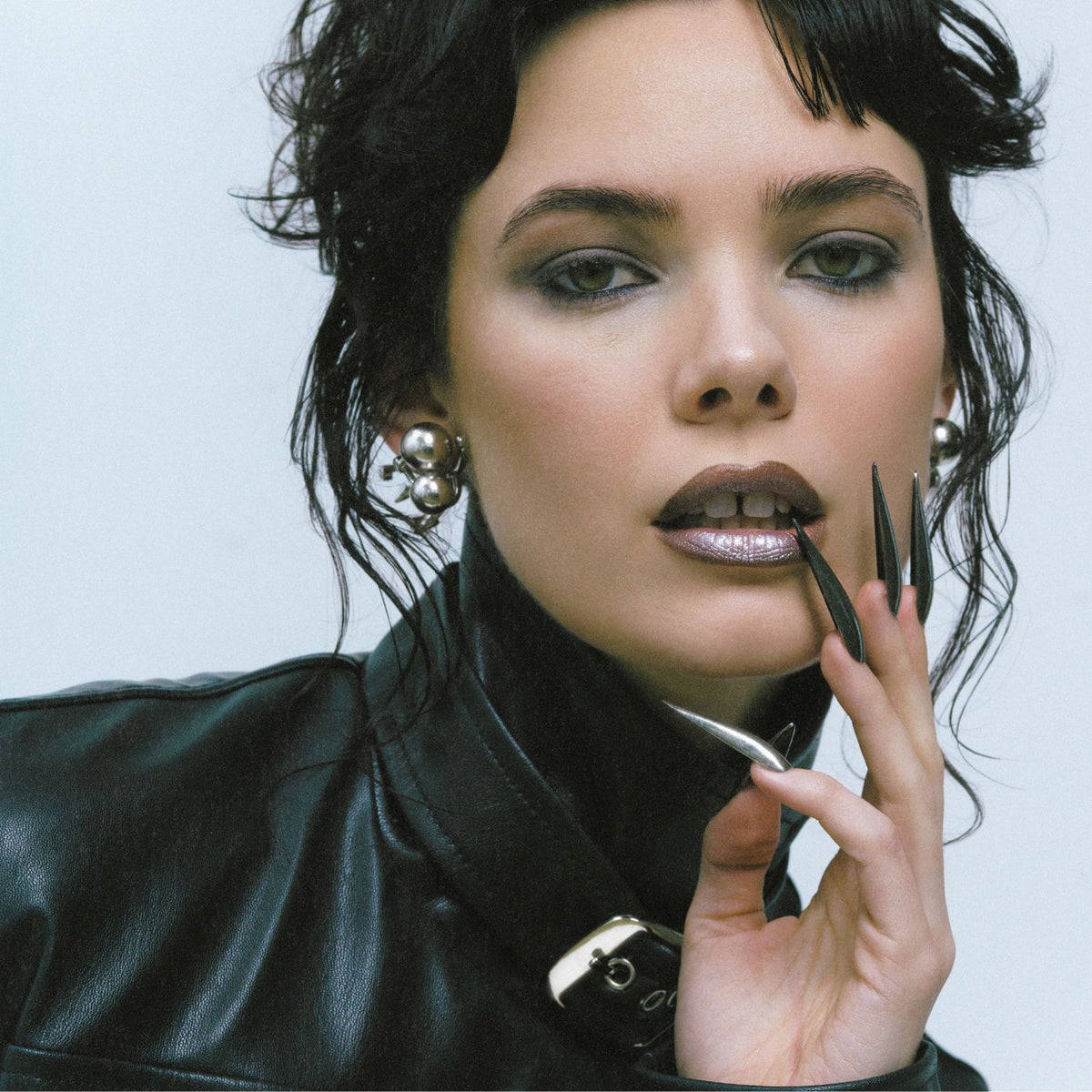 A woman with dark wavy hair wears JAREDJAMIN Jewelry Online Silver Uptown Orbita Clip On Earrings and a black leather jacket. She has metallic makeup, brown lipstick, and long black-and-silver pointed nails, gazing directly at the camera.