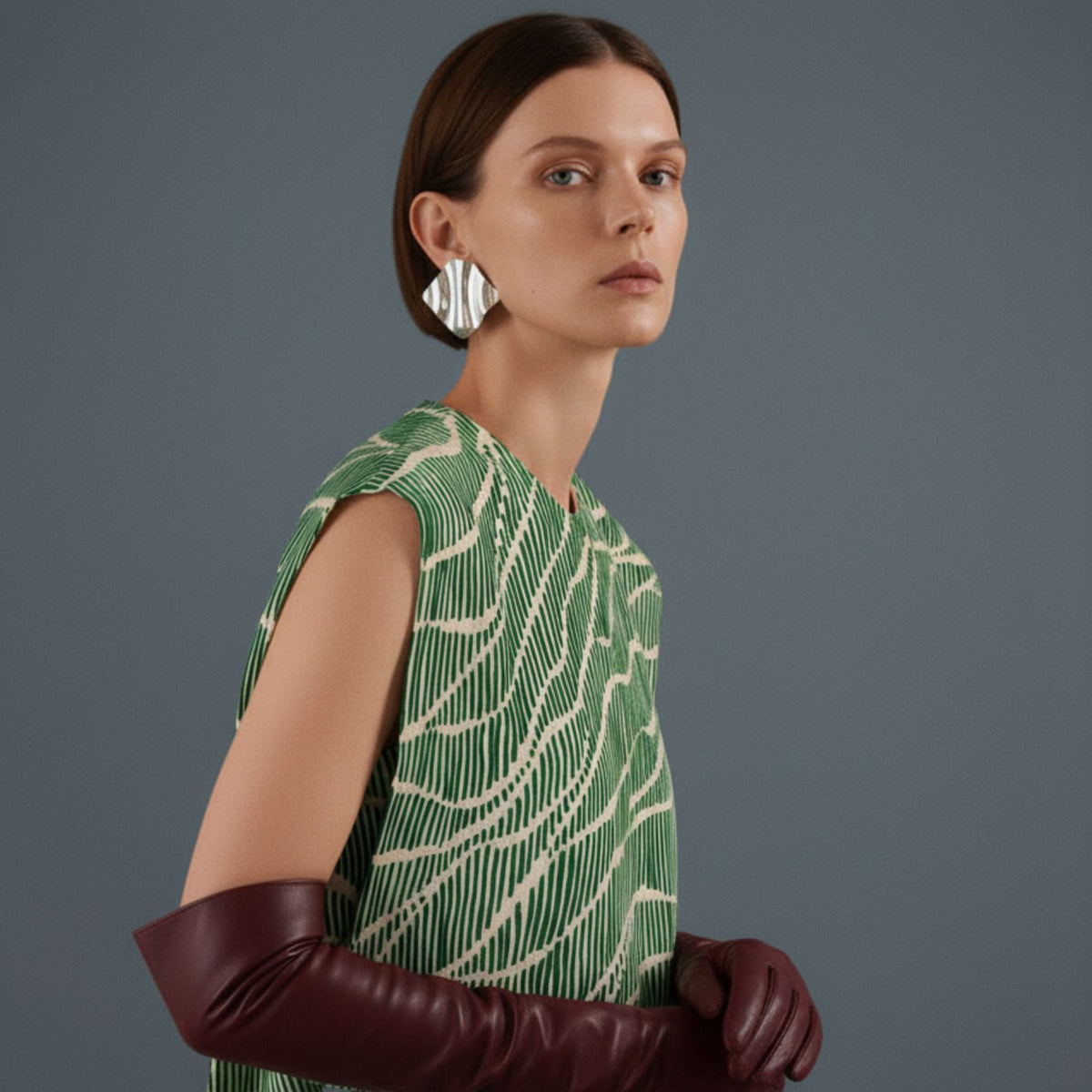 Un woman avec straight brown hair wears a sleeveless green et white patterned dress, long burgundy leather gloves, et the Whitney Wave boucles d'oreilles par JAREDJAMIN Bijoux en Ligne. She calmly poses against a plain gray background, looking toward the camera.
