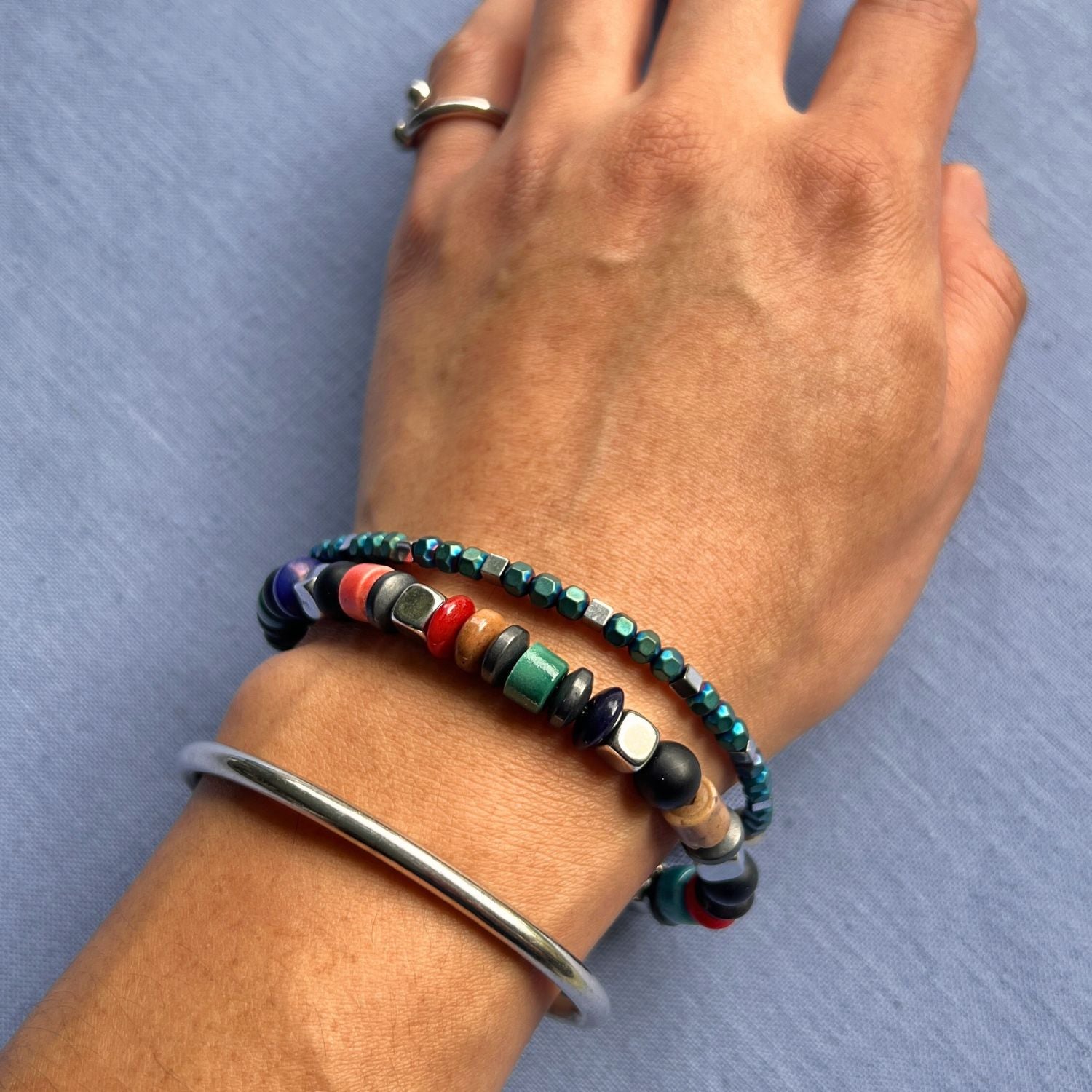 The Sloan Square Stretch Bracelet Set by JAREDJAMIN Jewelry Online features colorful round and square teal, red, green, black onyx gemstone, and silver beads with a red knot and metallic tag for a chic, eclectic style.