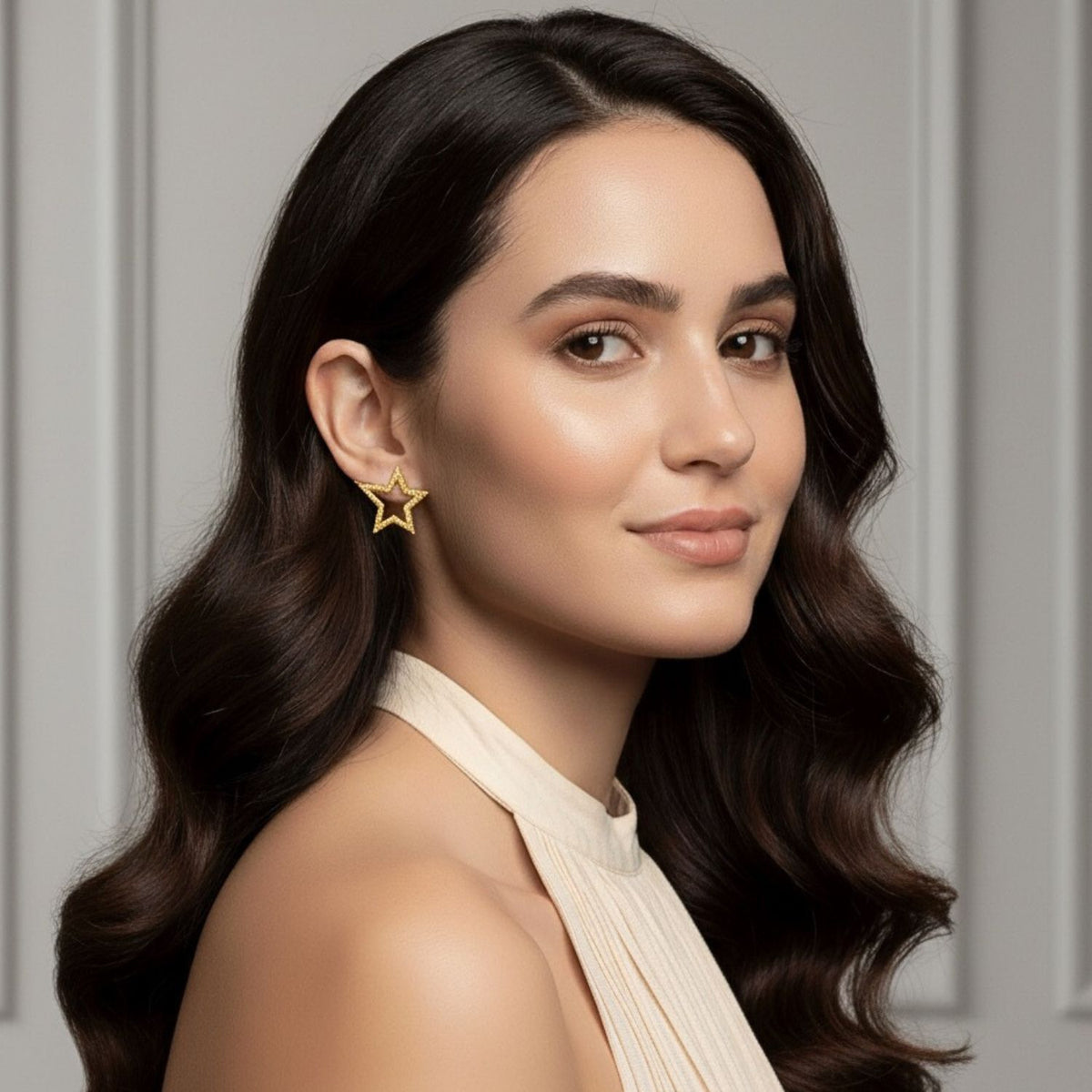 Un woman avec long, wavy dark brown hair et glowing skin smiles softly, wearing a sleeveless cream top et or Shooting Star Dust puce Post boucles d'oreilles Sm par JAREDJAMIN Bijoux en Ligne, against élégant light gray paneled walls.