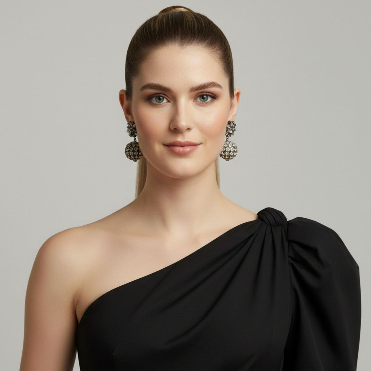 Un fair-skinned woman avec blue eyes et brown hair dans un high ponytail wears a sleek black one-shoulder dress et JAREDJAMIN Bijoux en Ligne’s Sparkling Bloom boucles d'oreilles. She has subtle makeup, et the background is light gray.