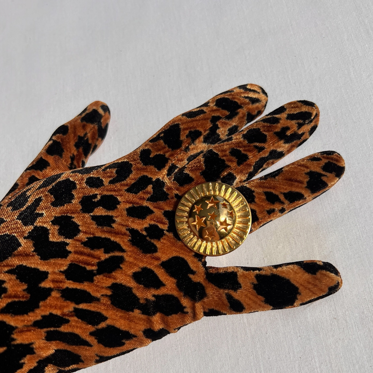 A hand in a leopard print glove elegantly displays the JAREDJAMIN Jewelry Online Starburst Statement Ring against a white background. Its gold, round design with embossed stars and ridged edges shines with celestial elegance, contrasting beautifully with the bold animal pattern.