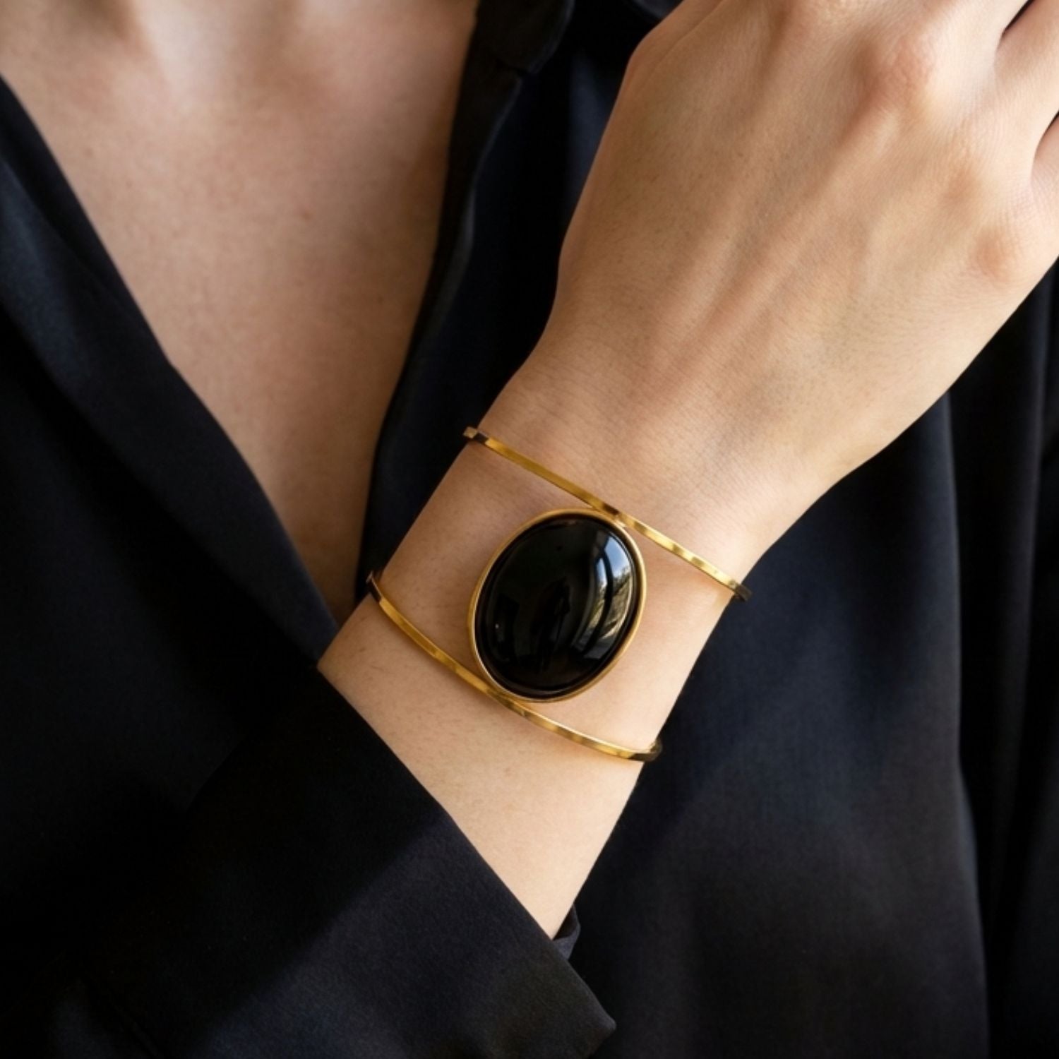A person in a black long-sleeve shirt wears the JAREDJAMIN Jewelry Online Stella Cuff In Black Agate: an 18K gold-plated brass bracelet with a glossy oval black stone and two slim gold bands elegantly encircling the wrist.