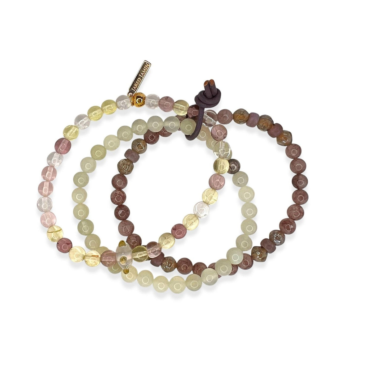 The Sunset Quarry Bracelet Set by JAREDJAMIN Jewelry Online features three overlapping bracelets with earthy-toned round beads—pale yellow, light brown, and reddish-brown—plus agate accents, a metal charm, and a brown knot closure.
