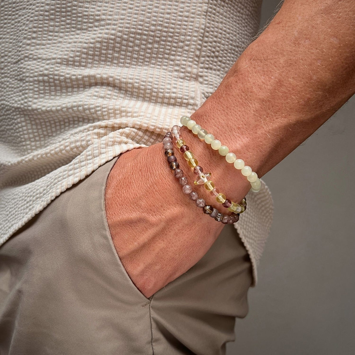 A close-up shows a hand in tan pants pocket, wearing the JAREDJAMIN Jewelry Online Sunset Quarry Bracelet Set with light green, transparent yellow, and pinkish-brown beads. The person also wears a textured off-white shirt.